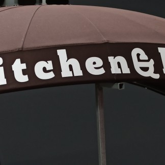 Front view — domed/eyebrow canopy on frame; white “Kitchen & Bath” lettering on brown fabric (prev. employer, lettering applied by Sunwell)